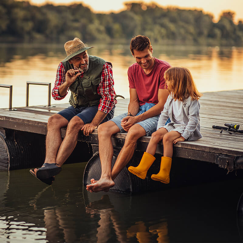 Grandpa sitting on a dock by the water with his son and granddaughter