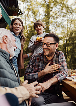 A grandfather laughing and preparing to eat outside at a picnic table with his son, daughter-in-law, and grandchild