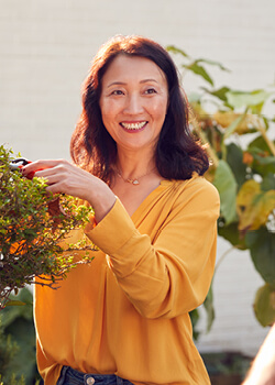 a woman with brown shoulder-length hair wearing a bright yellow sweater smiling and tending to a flower bush
